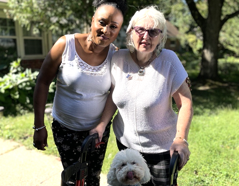 2 woman stand outside with a small white dog.