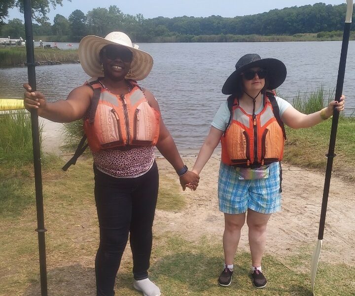 Two women on the bank of a river holding oars to kayak.